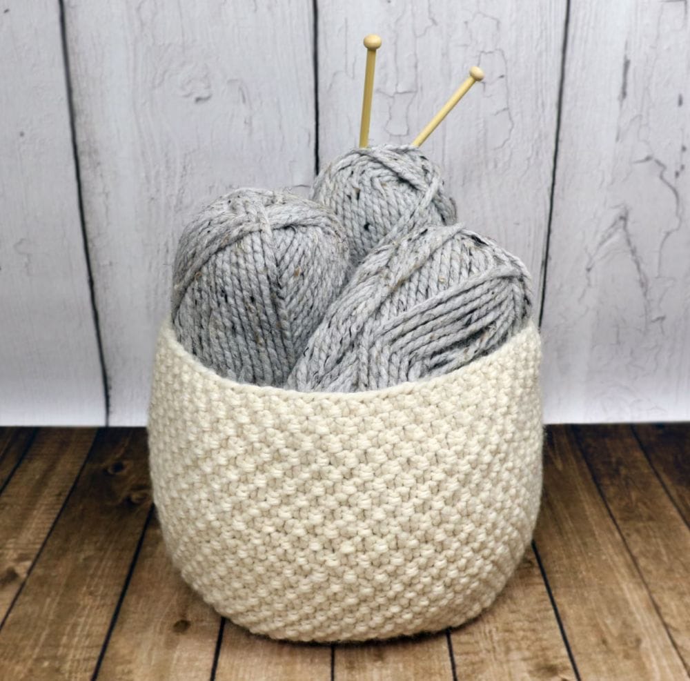 Oodles Basket by KnitAndCrochetEvrAft in Cream colored yarn holding gray skeins of yarn with wooden knitting needles sticking out from the skein
